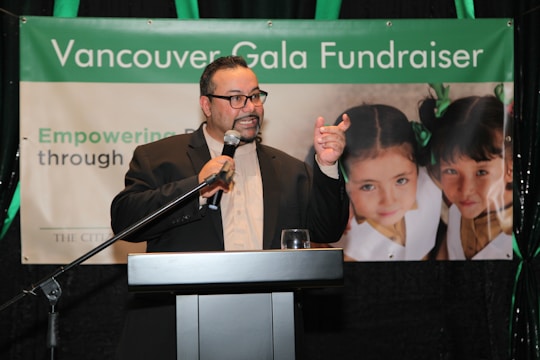 A person in a suit is speaking at a podium holding a microphone, with a banner in the background that reads 'Vancouver Gala Fundraiser'. The banner also features two young girls with green bows in their hair.