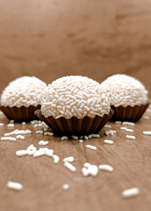 Three chocolate truffles are arranged on a light brown surface, each coated with white sprinkles and placed in a dark brown paper cup. The composition includes scattered white sprinkles around the truffles, conveying a homemade and rustic atmosphere.