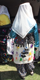 A person is shown from behind wearing a traditional outfit with a white headscarf. The outfit features colorful embroidery and tassels on a skirt or apron-like garment. The fabric is adorned with floral patterns in various bright colors, and the person appears to be part of a cultural or traditional gathering.