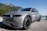 A modern, silver electric vehicle is parked on a gravel road, with a scenic backdrop of green mountains and a clear blue sky. The car has sleek lines and features branding text including 'Hyundai Ioniq 5' and 'All-electric' along its side.