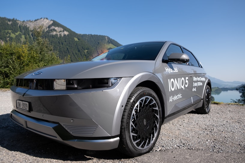 A modern, silver electric vehicle is parked on a gravel road, with a scenic backdrop of green mountains and a clear blue sky. The car has sleek lines and features branding text including 'Hyundai Ioniq 5' and 'All-electric' along its side.