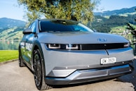 A sleek, modern electric vehicle is parked near a scenic lakeside with mountains in the background. The car has a smooth, futuristic design with a gray body and dark-tinted windows. There's a blue emblem on the front hood and a Swiss license plate. Surrounding the car, the lush greenery and clear sky add to the serene atmosphere.