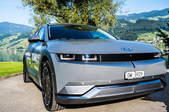 A sleek, modern electric vehicle is parked near a scenic lakeside with mountains in the background. The car has a smooth, futuristic design with a gray body and dark-tinted windows. There's a blue emblem on the front hood and a Swiss license plate. Surrounding the car, the lush greenery and clear sky add to the serene atmosphere.