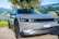 A sleek, modern electric vehicle is parked near a scenic lakeside with mountains in the background. The car has a smooth, futuristic design with a gray body and dark-tinted windows. There's a blue emblem on the front hood and a Swiss license plate. Surrounding the car, the lush greenery and clear sky add to the serene atmosphere.