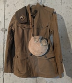 A vintage military-style brown jacket hangs against a rough, gray concrete wall. The jacket features buttoned pockets and shoulder straps. Draped over the shoulder is a matching shoulder belt. A dusty, round canteen with a metal top is attached to the jacket, giving a historical or retro vibe.
