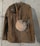 A vintage military-style brown jacket hangs against a rough, gray concrete wall. The jacket features buttoned pockets and shoulder straps. Draped over the shoulder is a matching shoulder belt. A dusty, round canteen with a metal top is attached to the jacket, giving a historical or retro vibe.