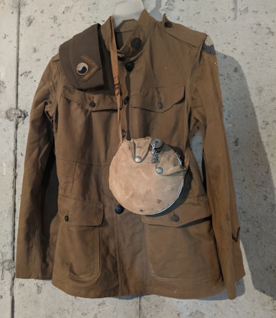 A vintage military-style brown jacket hangs against a rough, gray concrete wall. The jacket features buttoned pockets and shoulder straps. Draped over the shoulder is a matching shoulder belt. A dusty, round canteen with a metal top is attached to the jacket, giving a historical or retro vibe.