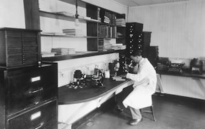 A man wearing a white lab coat is sitting on a stool and looking through a microscope in a laboratory. The laboratory is equipped with various scientific instruments, books, and filing cabinets. The surroundings are tidy with neatly arranged shelves filled with books and papers.