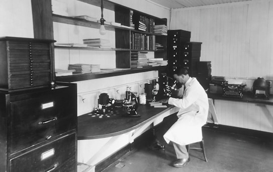 A man wearing a white lab coat is sitting on a stool and looking through a microscope in a laboratory. The laboratory is equipped with various scientific instruments, books, and filing cabinets. The surroundings are tidy with neatly arranged shelves filled with books and papers.