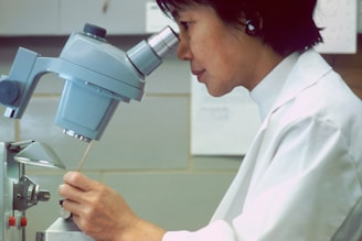 A person in a lab coat is closely observing something under a microscope, appearing focused and engaged in a scientific task. The background consists of a tiled wall and laboratory equipment.