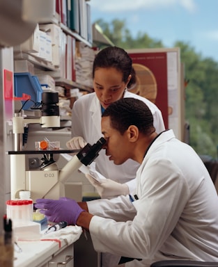 Two scientists in white lab coats are working in a laboratory, with one looking through a microscope and the other observing. The lab is equipped with various scientific equipment and supplies, including test tubes, a pipette, and laboratory storage shelves filled with containers and papers.