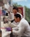 Two scientists in white lab coats are working in a laboratory, with one looking through a microscope and the other observing. The lab is equipped with various scientific equipment and supplies, including test tubes, a pipette, and laboratory storage shelves filled with containers and papers.