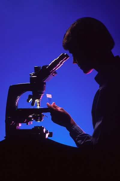 A profile of a female pathologist viewing a histological slide while sitting at a microscope. This dramatic shot has a blue background, with the microscope and the pathologist's features highlighted with magenta. Light is seen passing through the microscope's condensor to the objectives.