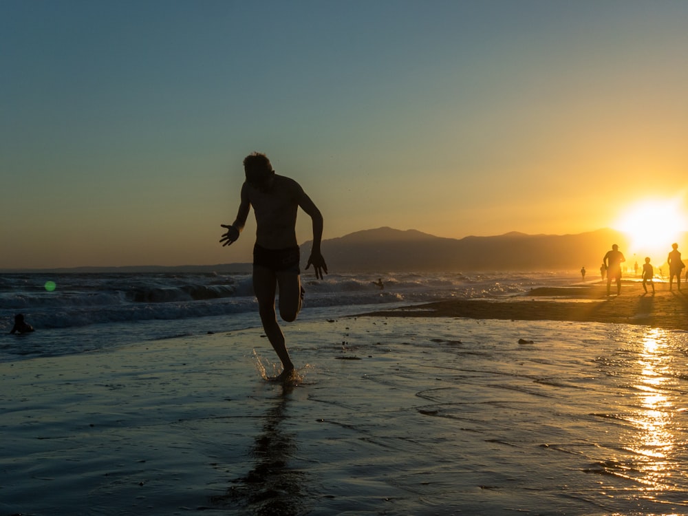 man in black tank top and blue shorts standing on beach during sunset