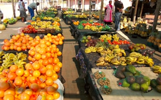 A vibrant fruit market stall displaying an array of colorful fruits including oranges, bananas, avocados, tomatoes, pineapples, and other assorted produce. The tables are laden with neatly arranged fruits, creating a vivid display. Several people are browsing the market, adding a sense of activity to the scene.