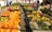 A vibrant fruit market stall displaying an array of colorful fruits including oranges, bananas, avocados, tomatoes, pineapples, and other assorted produce. The tables are laden with neatly arranged fruits, creating a vivid display. Several people are browsing the market, adding a sense of activity to the scene.