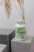 A bottle labeled 'Moringa Vital' is placed on top of geometric, gray textured blocks. The background features dried pampas grass adding a natural touch to the setting.