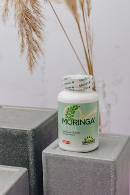A bottle labeled 'Moringa Vital' is placed on top of geometric, gray textured blocks. The background features dried pampas grass adding a natural touch to the setting.