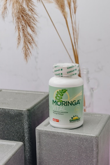 A bottle labeled 'Moringa Vital' is placed on top of geometric, gray textured blocks. The background features dried pampas grass adding a natural touch to the setting.