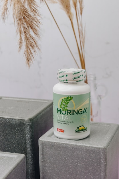 A bottle labeled 'Moringa Vital' is placed on top of geometric, gray textured blocks. The background features dried pampas grass adding a natural touch to the setting.