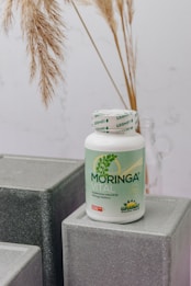 A bottle labeled 'Moringa Vital' is placed on top of geometric, gray textured blocks. The background features dried pampas grass adding a natural touch to the setting.