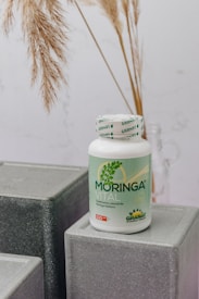 A bottle labeled 'Moringa Vital' is placed on top of geometric, gray textured blocks. The background features dried pampas grass adding a natural touch to the setting.