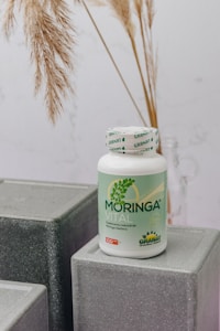 A bottle labeled 'Moringa Vital' is placed on top of geometric, gray textured blocks. The background features dried pampas grass adding a natural touch to the setting.