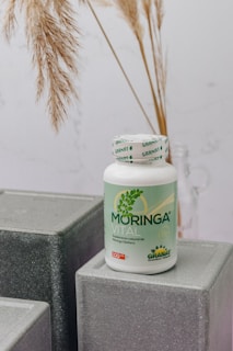A bottle labeled 'Moringa Vital' is placed on top of geometric, gray textured blocks. The background features dried pampas grass adding a natural touch to the setting.