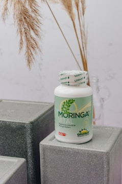 A bottle labeled 'Moringa Vital' is placed on top of geometric, gray textured blocks. The background features dried pampas grass adding a natural touch to the setting.