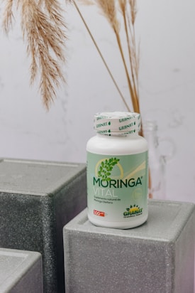 A bottle labeled 'Moringa Vital' is placed on top of geometric, gray textured blocks. The background features dried pampas grass adding a natural touch to the setting.