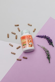 A bottle labeled 'REDU GRAN PLUS' is surrounded by several brown capsules, with two sprigs of lavender placed on a purple and off-white geometrically laid-out background, suggesting a health or wellness theme.
