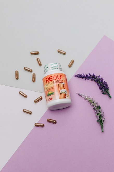 A bottle labeled 'REDU GRAN PLUS' is surrounded by several brown capsules, with two sprigs of lavender placed on a purple and off-white geometrically laid-out background, suggesting a health or wellness theme.