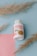 A white bottle with a red and orange label is positioned at the center. The label reads 'FENO GRAN PLUS' with additional text related to the product's natural ingredients. The bottle is surrounded by decorative dried grass stems, resting on a pastel blue background with a pink paper accent.