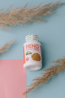 A white bottle with a red and orange label is positioned at the center. The label reads 'FENO GRAN PLUS' with additional text related to the product's natural ingredients. The bottle is surrounded by decorative dried grass stems, resting on a pastel blue background with a pink paper accent.