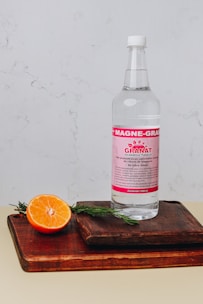 A bottle of mineral supplement labeled 'Magne-Granat' sits on a wooden cutting board. Beside it, a halved orange and sprigs of rosemary add a touch of natural freshness. The background features a light, textured wall.