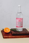 A bottle of mineral supplement labeled 'Magne-Granat' sits on a wooden cutting board. Beside it, a halved orange and sprigs of rosemary add a touch of natural freshness. The background features a light, textured wall.