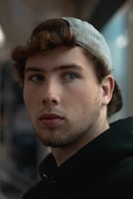 A young person with short hair is wearing a gray cap backwards and a black hoodie, gazing slightly to the side. The lighting is subdued and soft, creating a moody atmosphere.