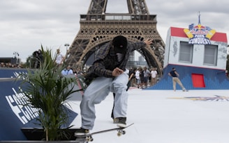 A skateboarder performs a trick in front of the Eiffel Tower. The scene includes a skating ramp with 'Red Bull Paris Conquest' signage. There are several people in the background watching the event. The skateboarder is wearing a plaid shirt and light blue jeans.