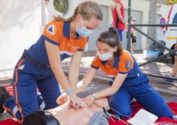 Two individuals wearing medical uniforms and face masks are performing CPR on a training mannequin. They are on their knees, focused on administering chest compressions within an outdoor setting. The scene suggests a training or emergency response demonstration.