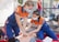 Two individuals wearing medical uniforms and face masks are performing CPR on a training mannequin. They are on their knees, focused on administering chest compressions within an outdoor setting. The scene suggests a training or emergency response demonstration.
