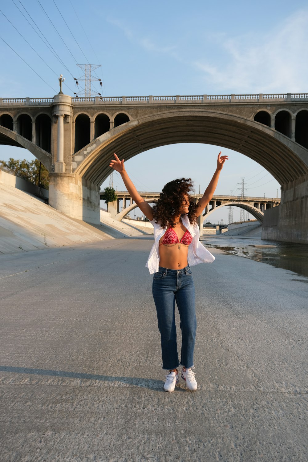 Mujer con crop top blanco y jeans de mezclilla azul de pie en un puente de concreto gris durante el día