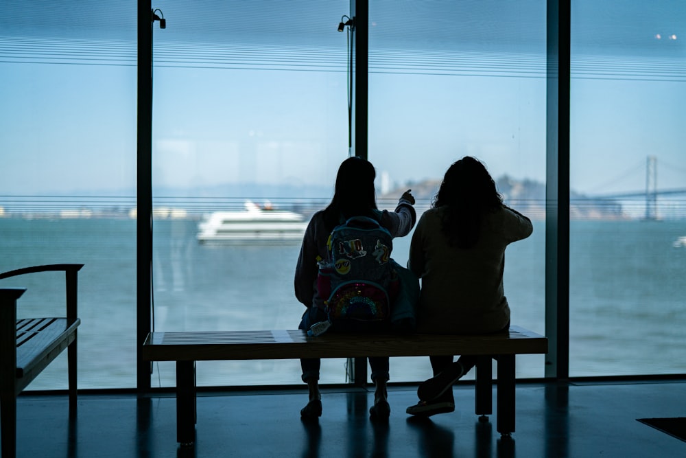 2 women sitting on brown wooden chair looking at the window during daytime