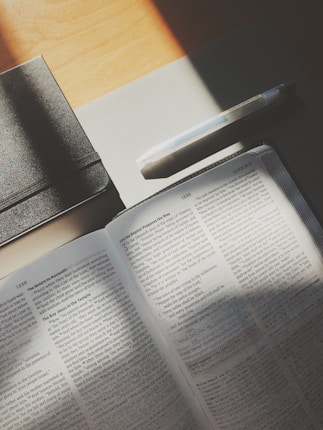An open book lies on a table, with sunlight streaming onto its pages. A black notebook and a white pen are placed nearby, casting gentle shadows.