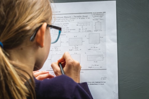 A person with long hair, wearing glasses, is focused on a worksheet with math problems related to measuring unknown perimeters. The individual is using a pencil to write or solve problems on the paper, which has various geometric shapes and equations printed on it.