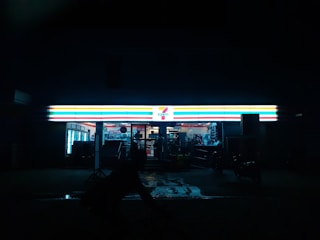 A convenience store with a brightly illuminated sign stands out against the dark night. The interior is faintly visible, displaying shelves and products. A silhouette of a person or object is seen in front of the store.
