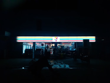 A convenience store with a brightly illuminated sign stands out against the dark night. The interior is faintly visible, displaying shelves and products. A silhouette of a person or object is seen in front of the store.