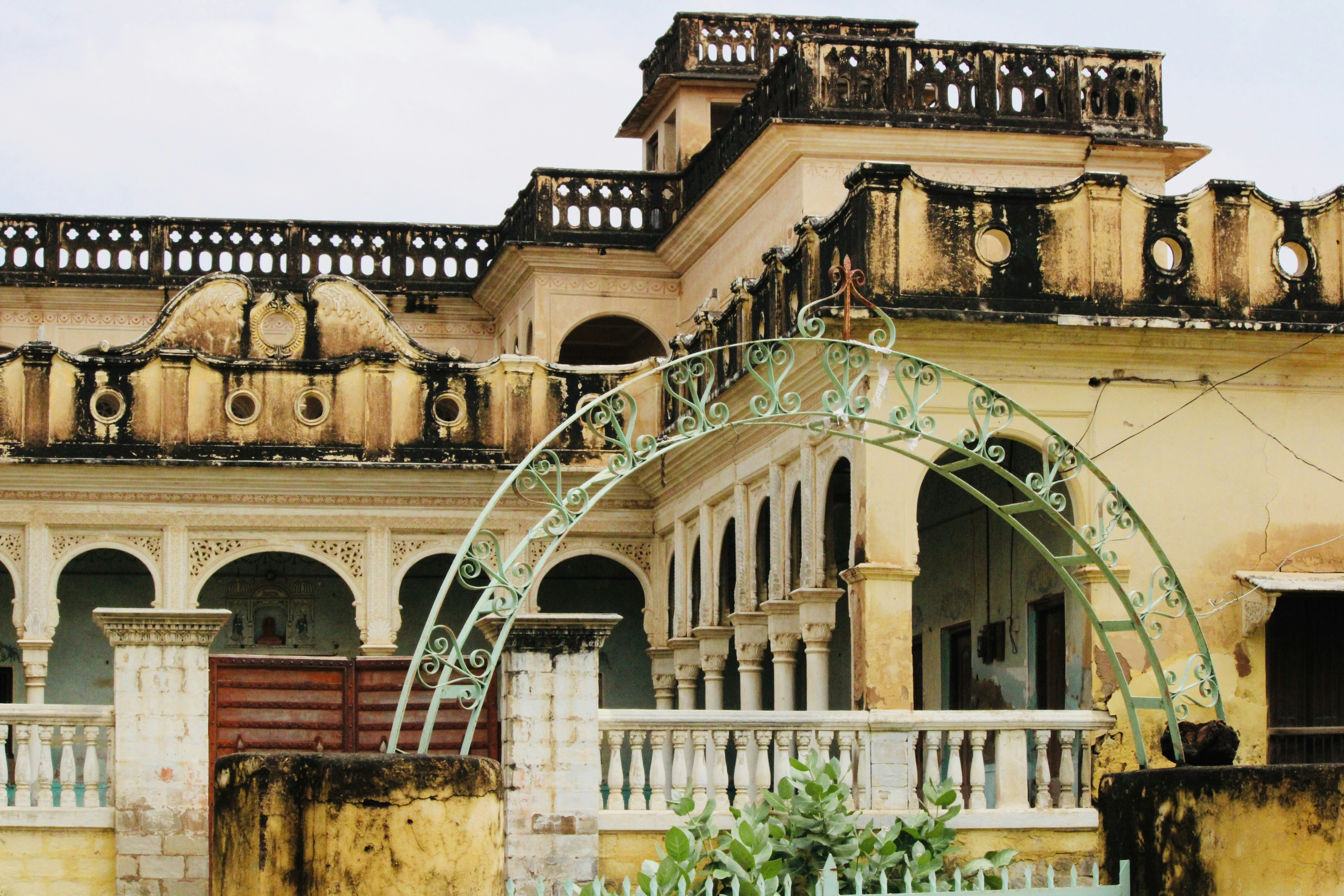 Photograph of a weathered yellow veranda framed by a circular verdigris arch with ornate railings. The composition emphasizes architectural decay and decorative metalwork.