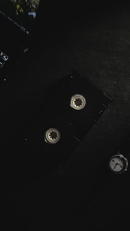 A vintage video cassette is placed on a dark surface, alongside part of a laptop keyboard in the top left corner and a watch in the bottom right. The lighting is dim and creates a moody atmosphere.
