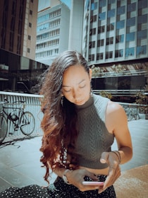 A woman with long, wavy hair is sitting outdoors in an urban setting, looking down at her smartphone. Tall buildings surround her, and bicycles are parked nearby. She is wearing a sleeveless cable-knit top and a polka dot skirt, with sunlight casting shadows on the scene.