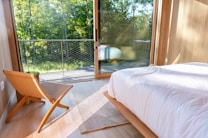 A modern bedroom features a large glass door opening to a balcony with a view of lush green trees. The room has a minimalistic design with wooden elements, including a woven chair and light wooden flooring. A white bedspread covers the neatly made bed, and sunlight filters through the glass, casting soft shadows on the floor.